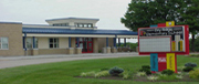 A single-story building with a flat roof and large windows, featuring a colorful sign in front, surrounded by green grass and shrubs.