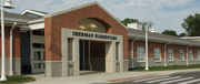 The exterior of a brick building with a sign that reads 'Sherman Elementary' above the entrance, featuring large windows and a landscaped area.