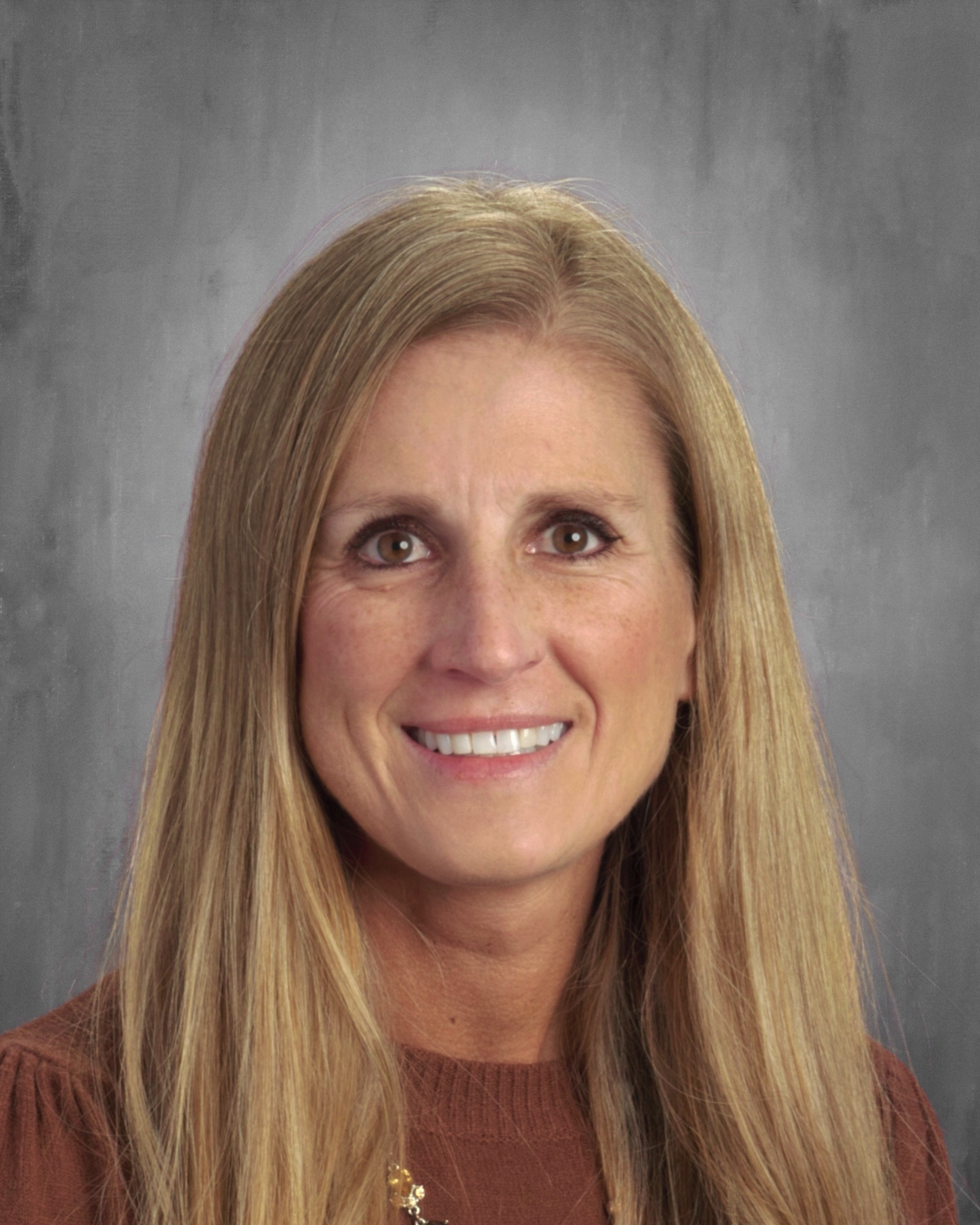 A smiling woman with long, straight blonde hair, wearing a brown sweater, is posed against a gray background.