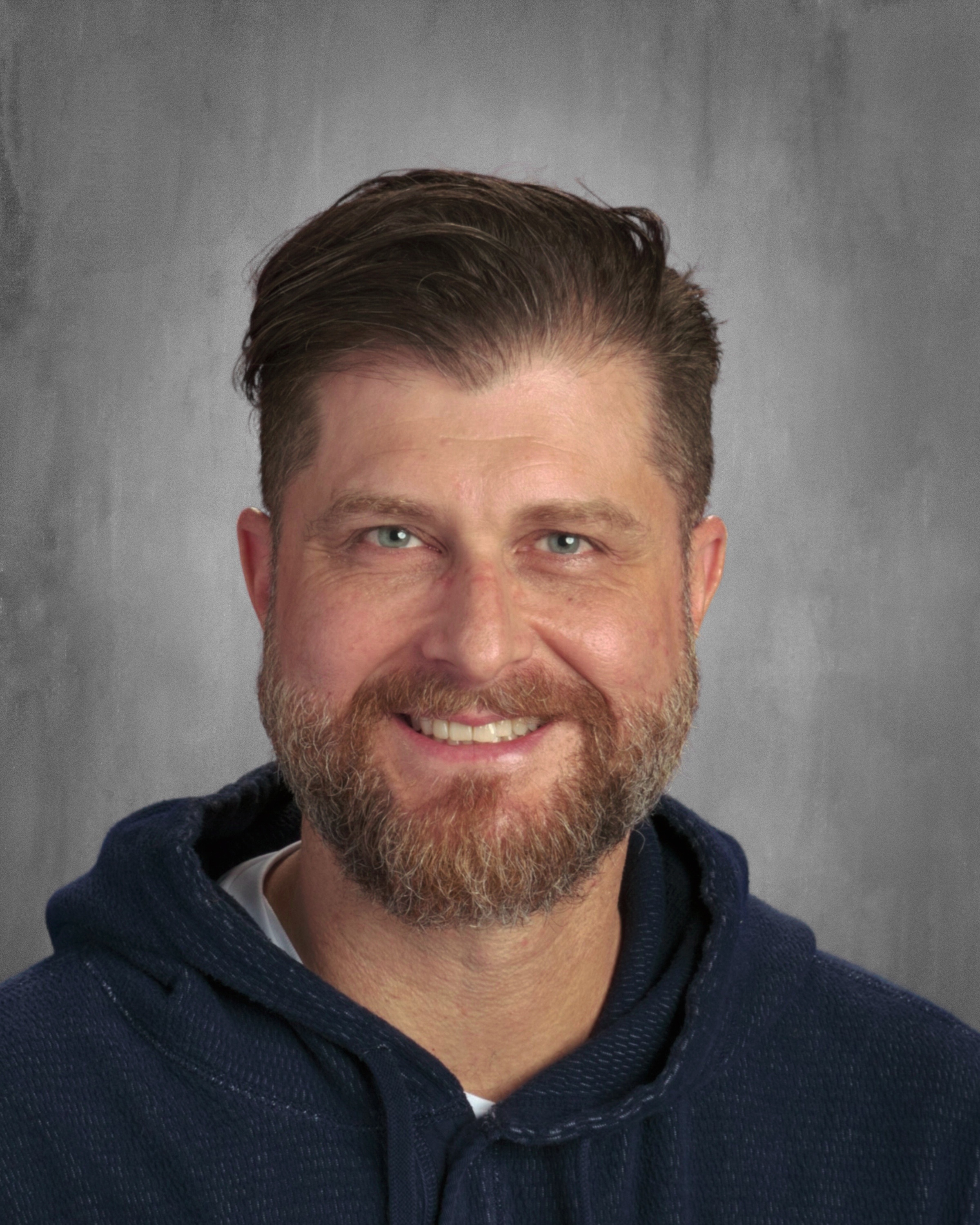 A smiling man with a beard and short hair, wearing a dark hoodie, against a gray background.