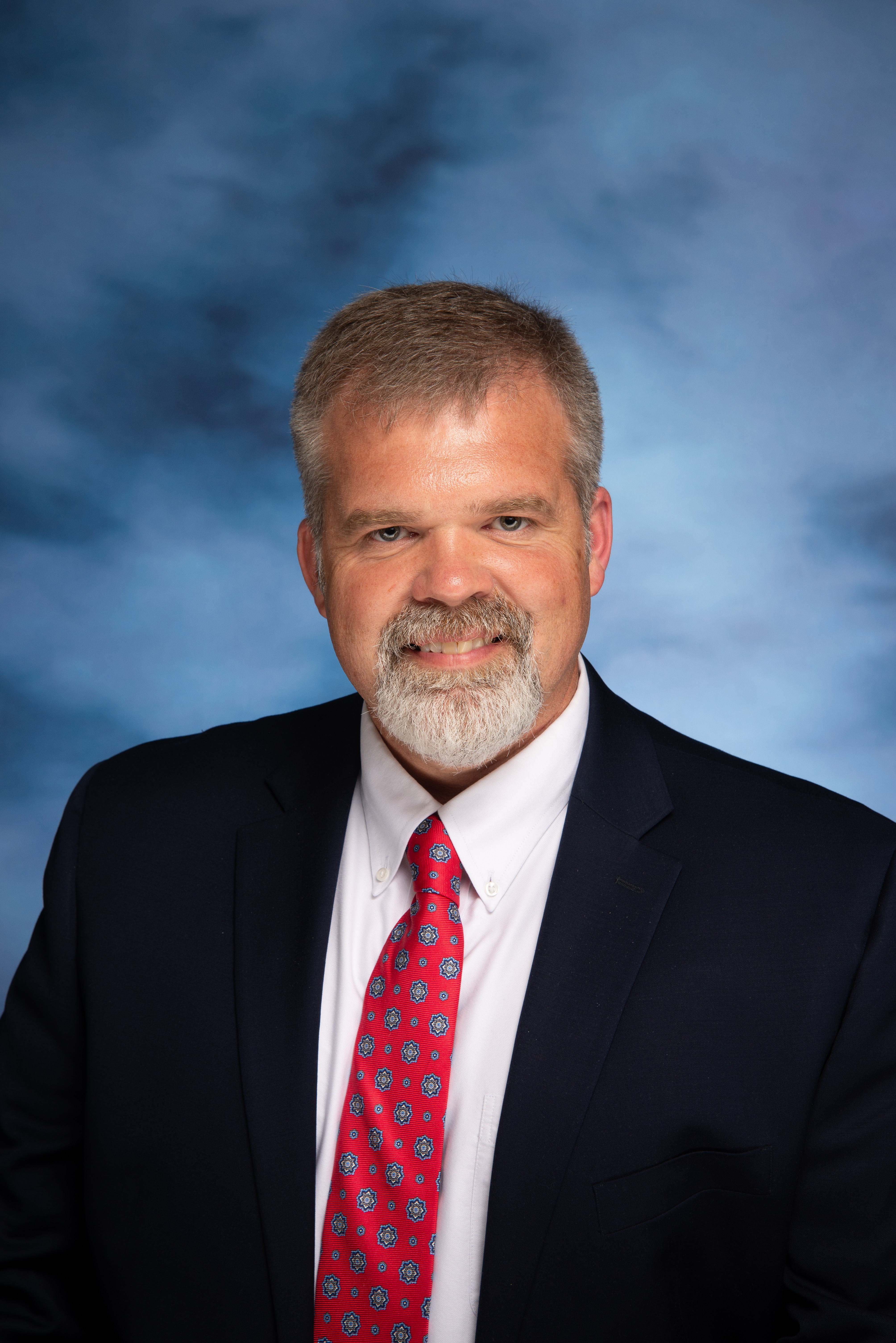 A man with a beard and short hair, wearing a dark suit and a white shirt with a red and pink patterned tie, smiles against a blue background.