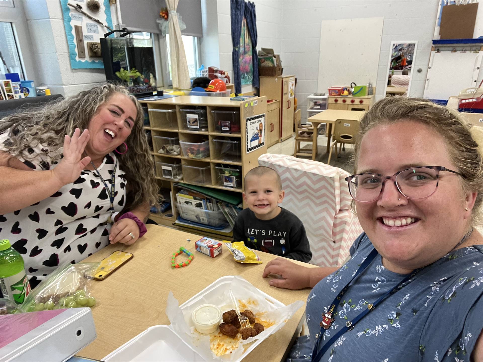 Three individuals, two adults and a child, are smiling and enjoying a meal together at a table in a classroom setting, with educational materials and furniture visible in the background.