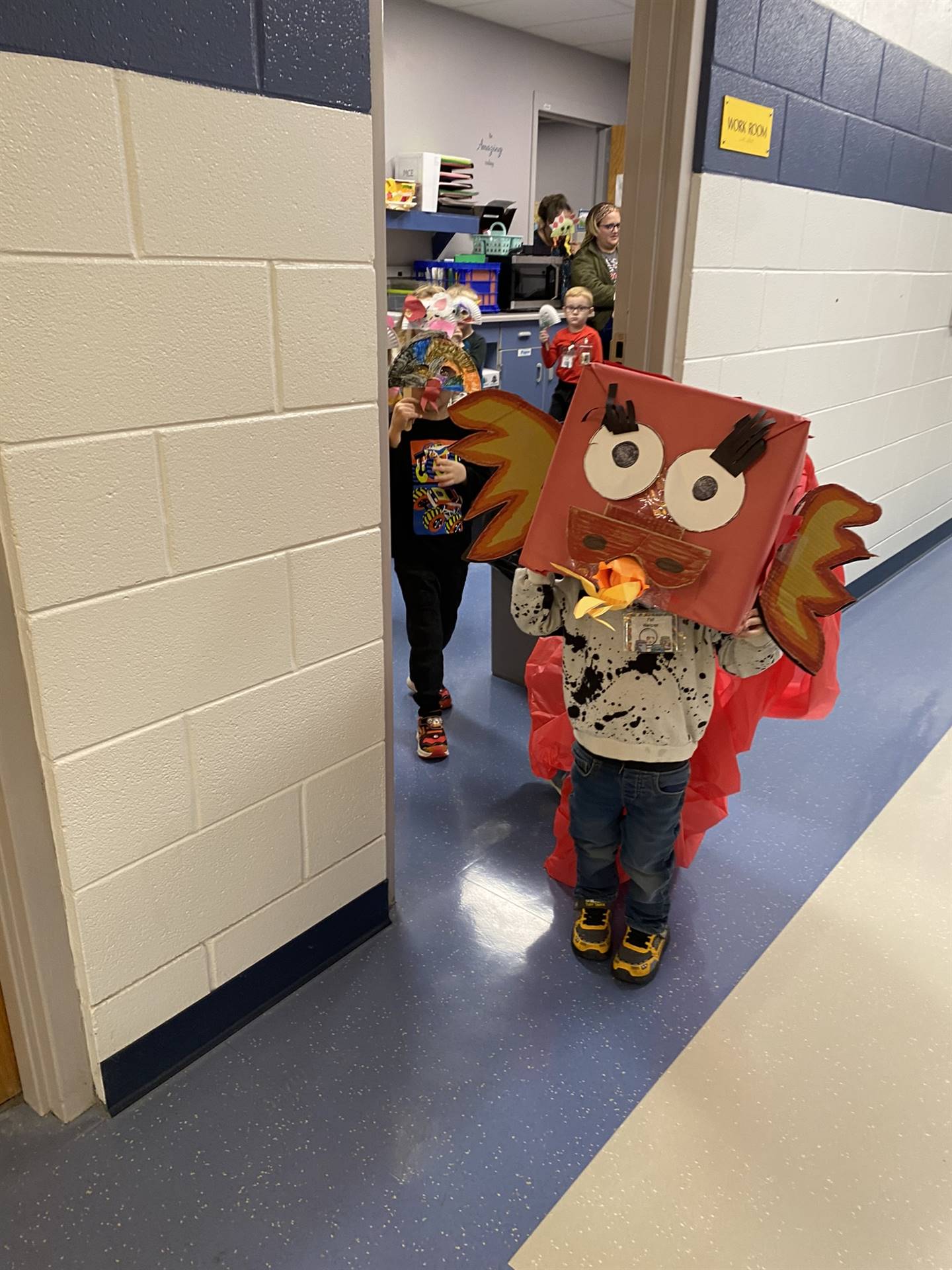 Children wearing colorful costumes, including a large dragon mask, walk through a school hallway decorated for an event.