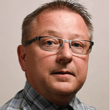 Headshot of a middle-aged man with short light brown and gray hair wearing rectangular glasses, a gray plaid button-down shirt, and a dark tie, looking at the camera against a neutral background.