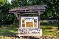 Wooden sign for Black Kettle North Park in Kansas, featuring green graphics and surrounded by lush trees.