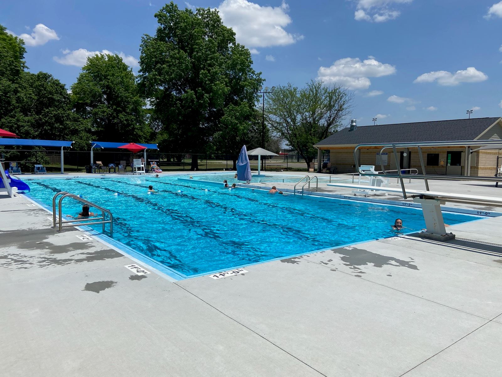 A sunny outdoor swimming pool with blue water, surrounded by trees and lounge chairs, featuring a diving board and umbrellas.