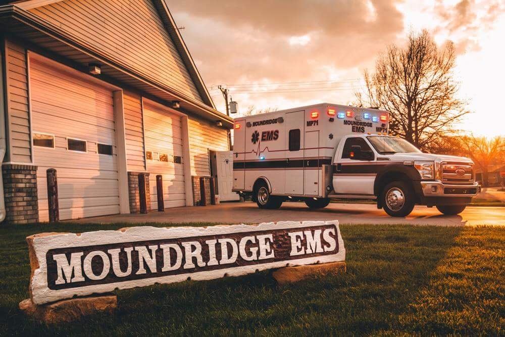 A Moundridge EMS ambulance parked in front of the station, illuminated by a sunset, with a sign reading "Moundridge EMS" in the foreground.