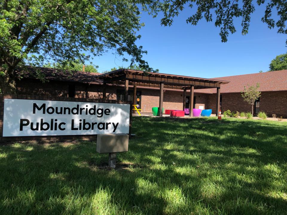 Moundridge Public Library building surrounded by green grass and colorful outdoor seating under a sunny blue sky.