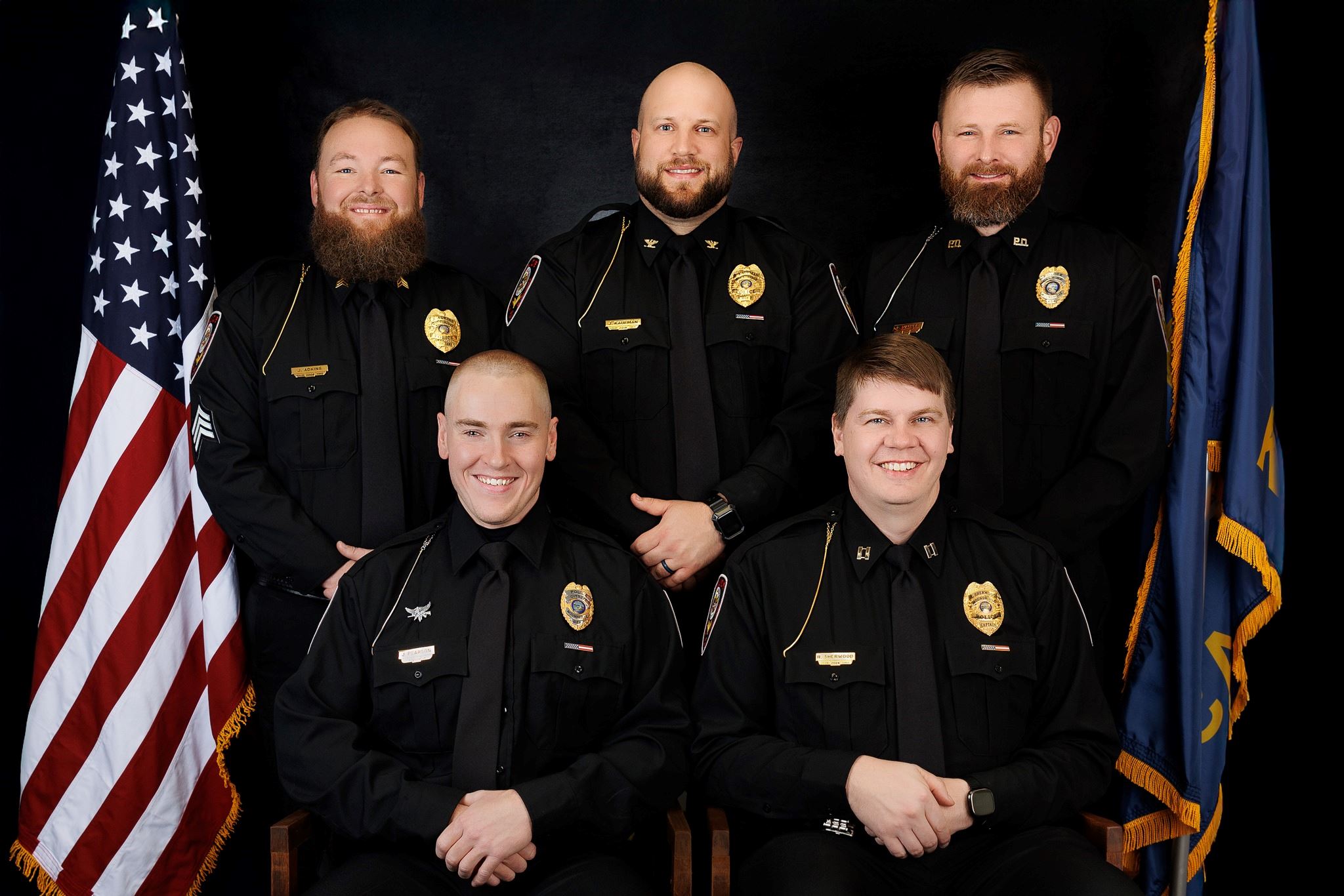 A group of six police officers in uniform, posed in front of a black backdrop, with an American flag and a blue flag displayed.