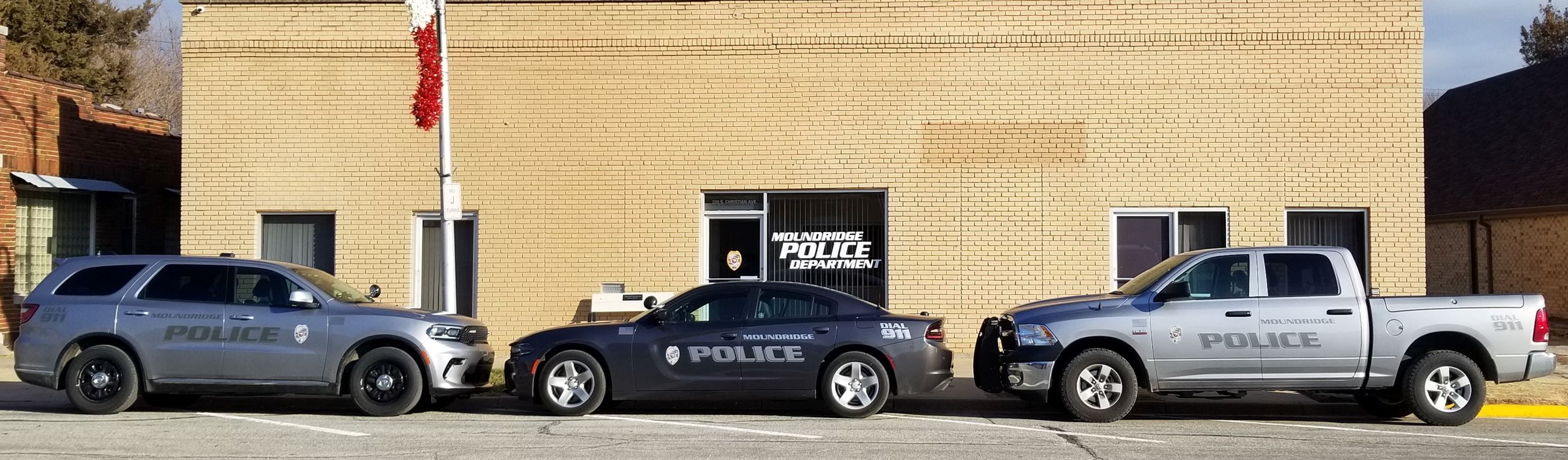 Three Moundridge police vehicles—an SUV, a sedan, and a pickup truck—parked outside the police department building.