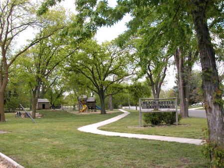 A scenic park with green trees, a winding path, and a sign for Black Kettle East Park, featuring playground equipment in the background.