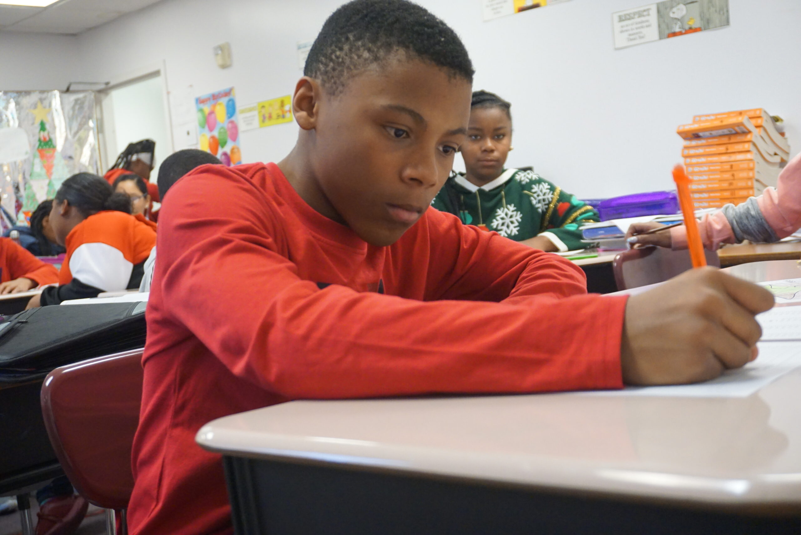 A young boy wearing a red shirt is focused on writing at his desk in a classroom setting.