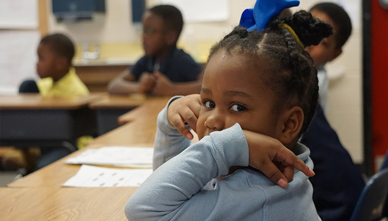 A young girl sitting at a classroom desk, focused on her work with books and stationery around her.
