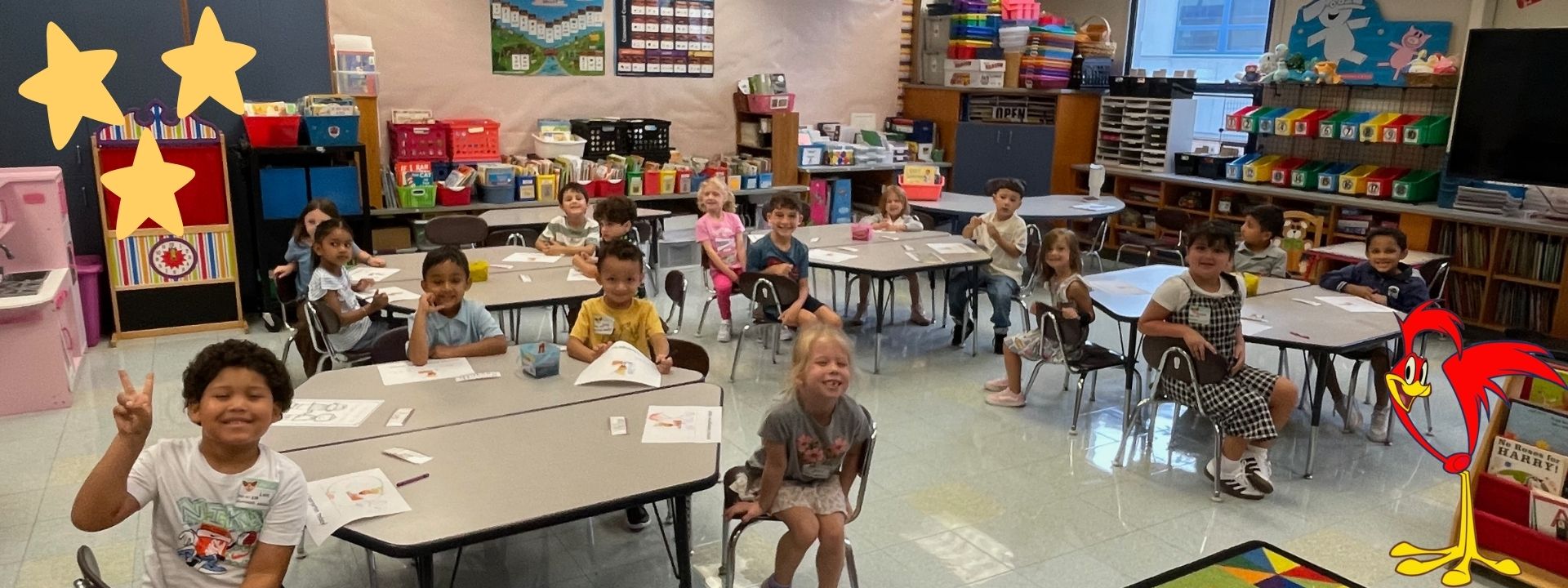 Classroom with students seated at tables. Some wear casual clothes. Colorful decorations and books on shelves.