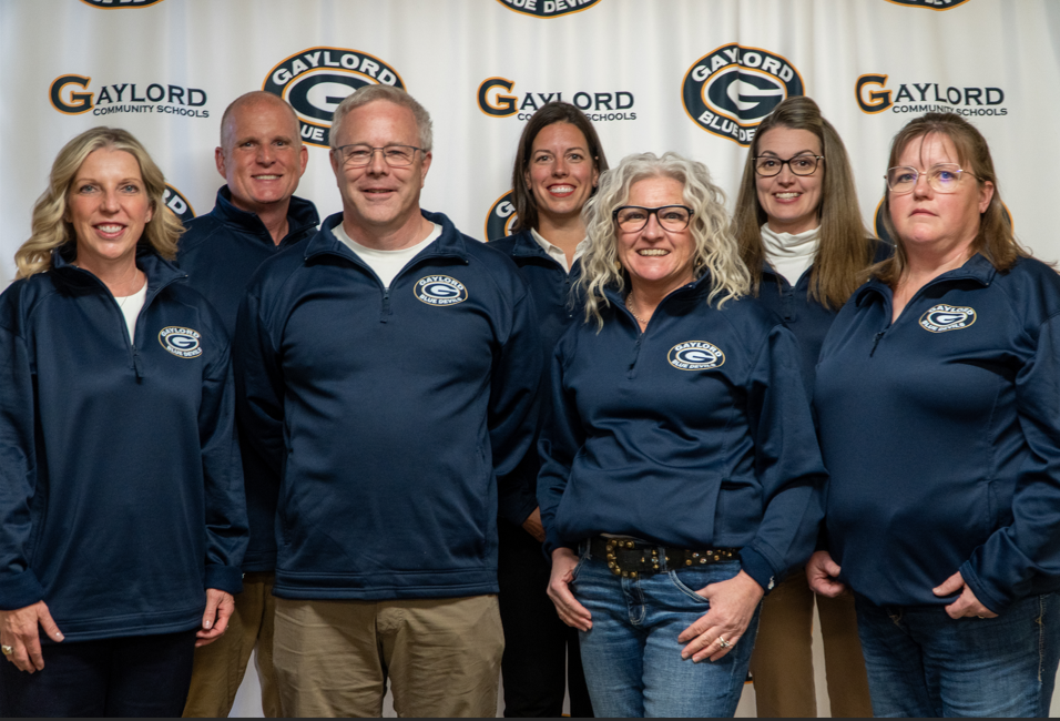 A group of seven individuals, wearing matching jackets, pose for a photo in front of a Gaylord Community Schools sign, with two seated in front and five standing behind them.