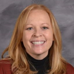 A smiling woman with long, wavy blonde hair, wearing a black top and a brown cardigan, poses for a portrait against a gray background.