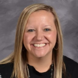 A smiling woman with long blonde hair, wearing a black shirt and necklace, poses for a portrait against a gray background.