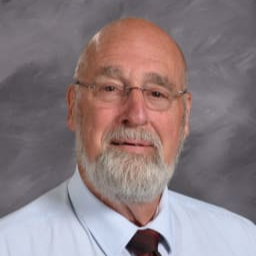 A senior man with glasses and a beard, wearing a light blue shirt and a dark tie, poses for a portrait against a gray background.
