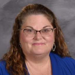 A woman with long, curly brown hair wearing glasses and a blue blouse, smiling at the camera against a gray background.