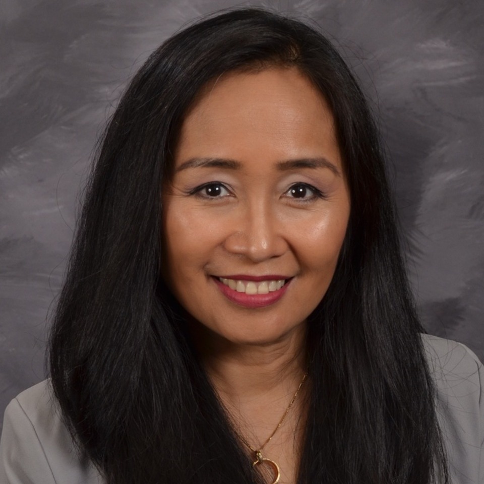 A woman with long black hair and a warm smile is wearing a light blazer, set against a softly blurred gray background.