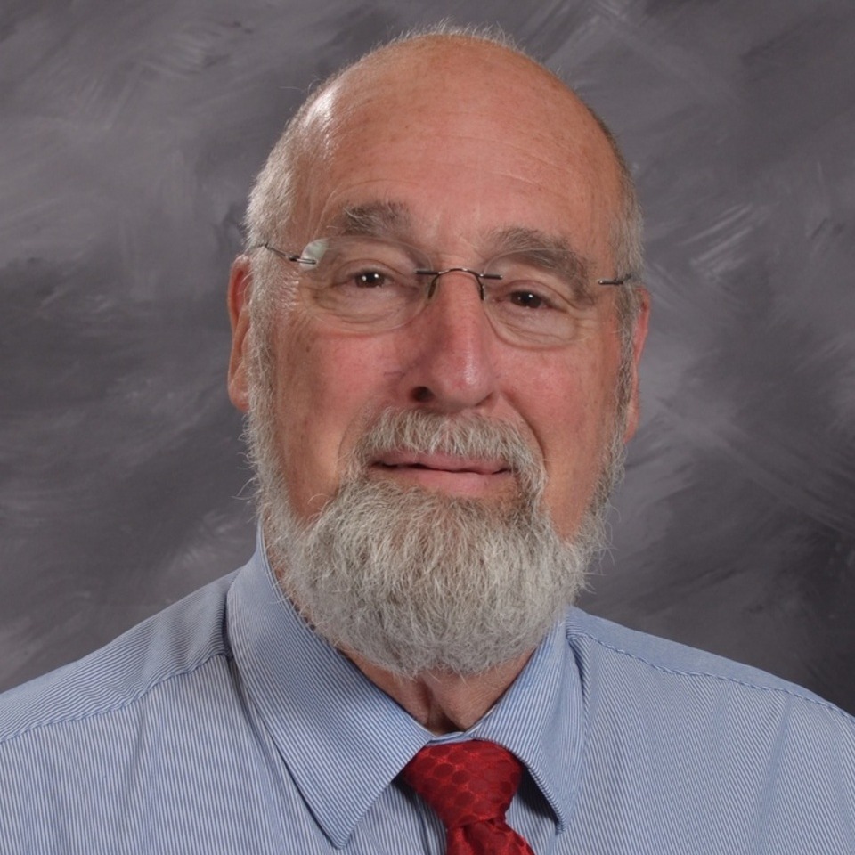 A senior man with glasses and a beard, wearing a light blue shirt and a dark tie, poses for a portrait against a gray background.