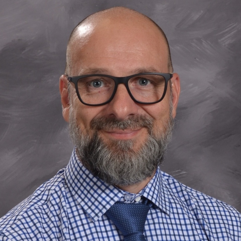 Bald man wearing glasses, blue checked shirt and tie, against a grey background