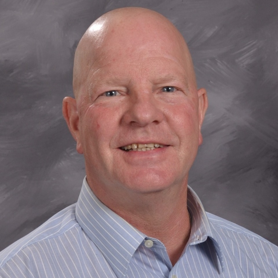 A man with no hair, smiling, wearing a light blue shirt, against a grey background