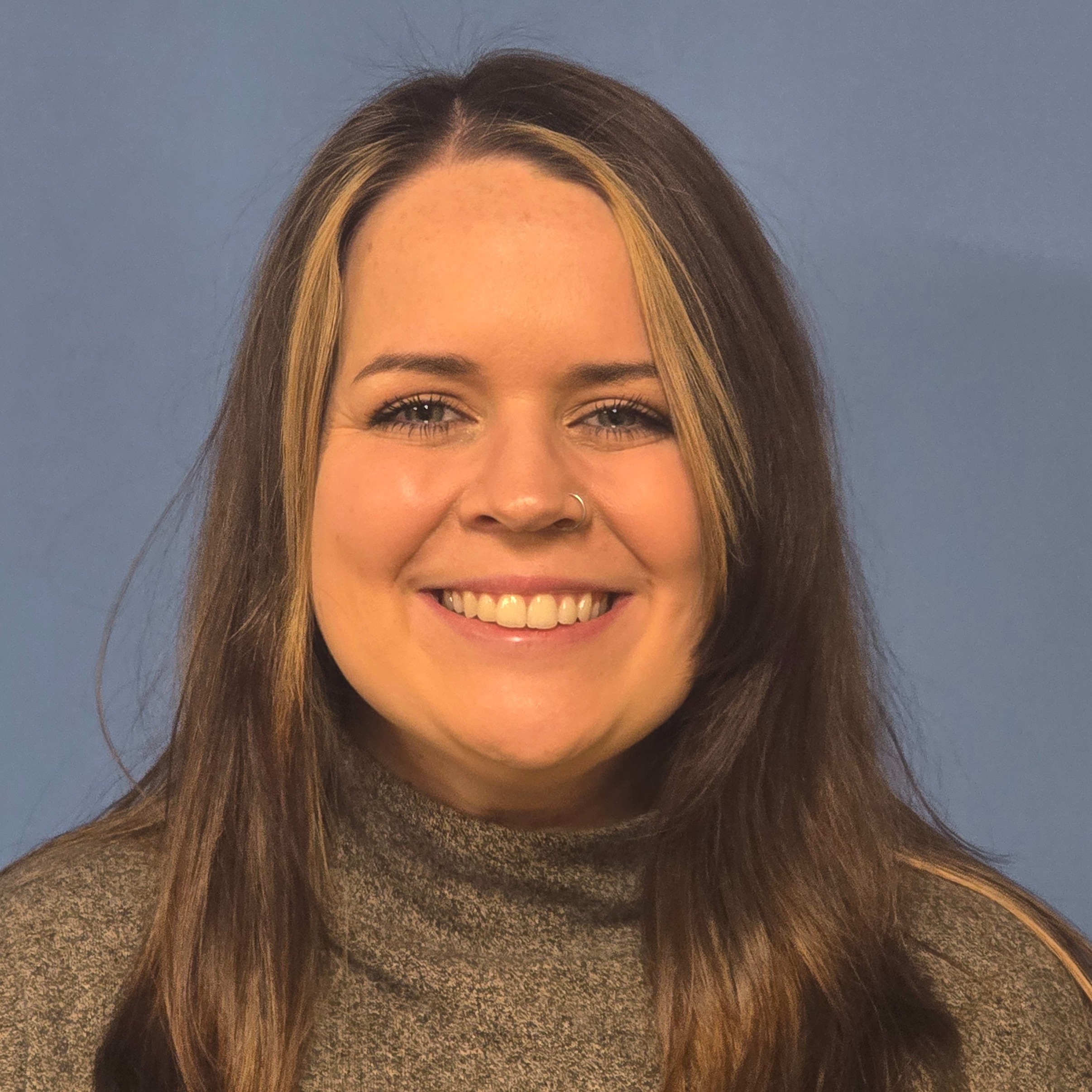 A smiling woman with long brown hair, wearing a a dark shirt, poses for a portrait against a blue background.
