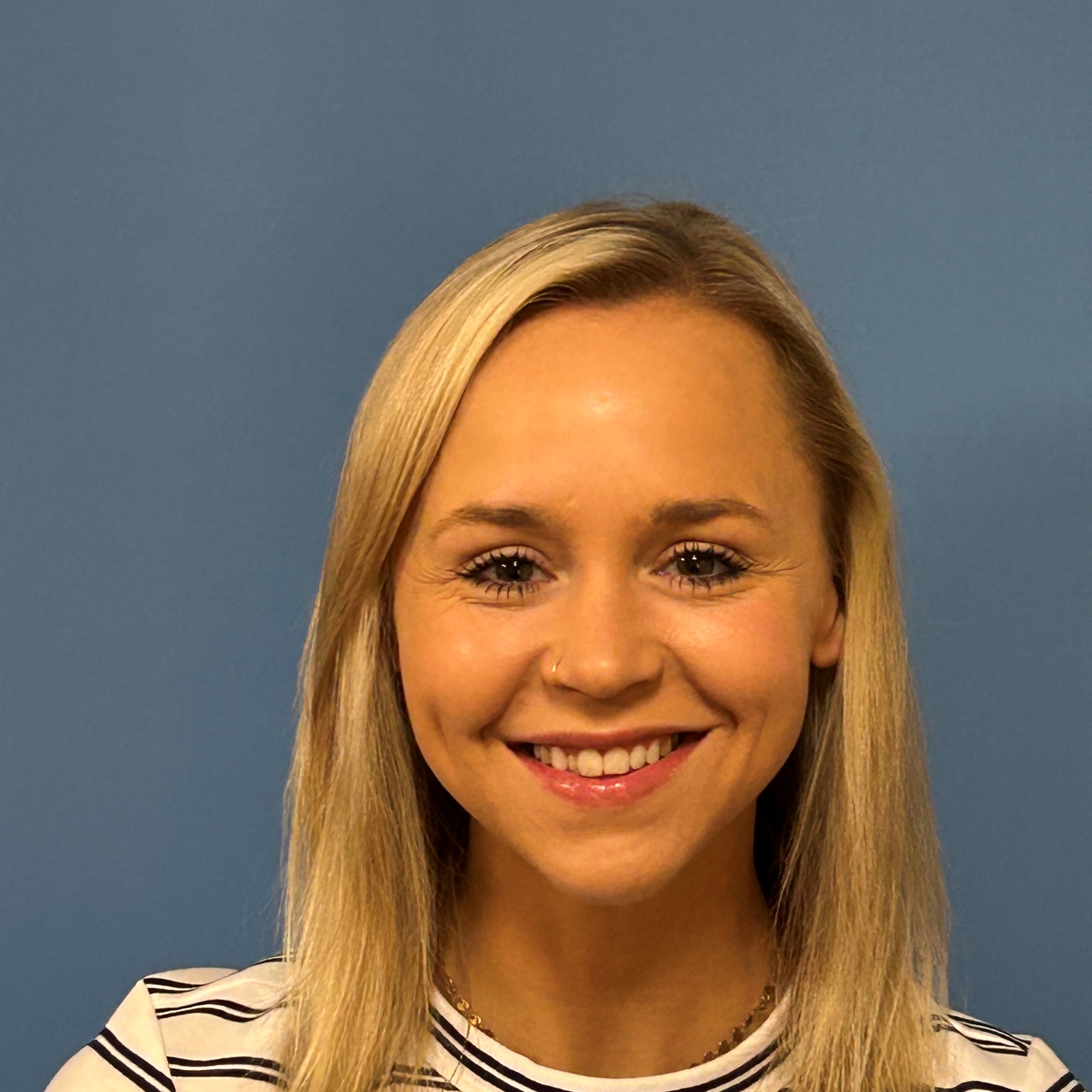 A smiling woman with long, blonde hair, wearing a stripe top, poses for a portrait against a gray background.