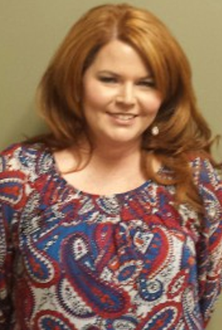 A woman with long, wavy red hair is smiling while wearing a colorful paisley-patterned blouse against a plain background.