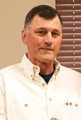 A man with short dark hair and a slight smile, wearing a light-colored button-up shirt, stands in front of a window with blinds.