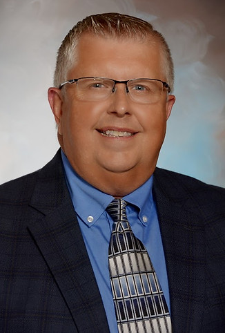 A smiling man with short, light-colored hair and glasses, wearing a blue dress shirt and a patterned tie, poses for a professional headshot against a soft, neutral background.