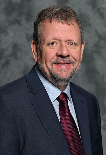 A middle-aged man with a beard and short hair, wearing a dark suit and a maroon tie, smiles against a gray background.