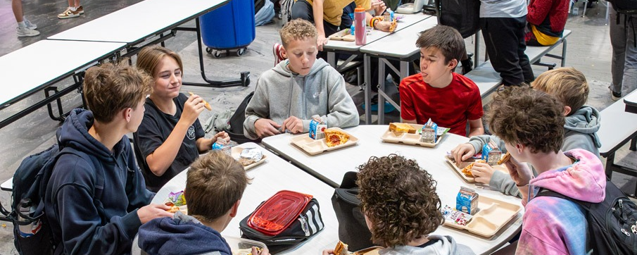 Middle school students eat lunch at round and rectangular cafeteria tables. A variety of foods and milk cartons are visible. Other students line up in the background. The scene is busy and social.