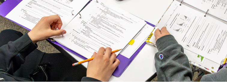 Two students working at a table with binders and worksheets; one holds a pencil over a page with text and graphs.