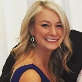 A smiling woman with long, wavy blonde hair wearing a blue dress and dangling earrings, posed against a light background.