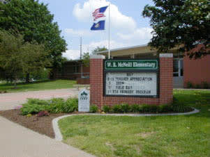 A view of the exterior of M. J. McNutt Elementary School, featuring a sign with information about the school and flags flying in the background.