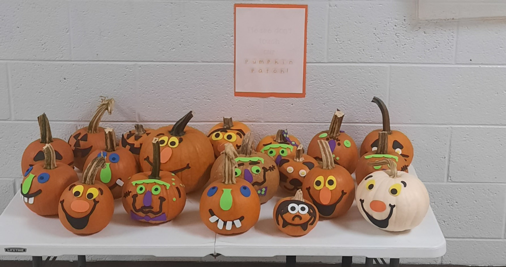 A table displays colorful, painted pumpkins with various smiling faces and silly expressions, alongside a sign reading "Please don’t touch our Pumpkin Patch!"