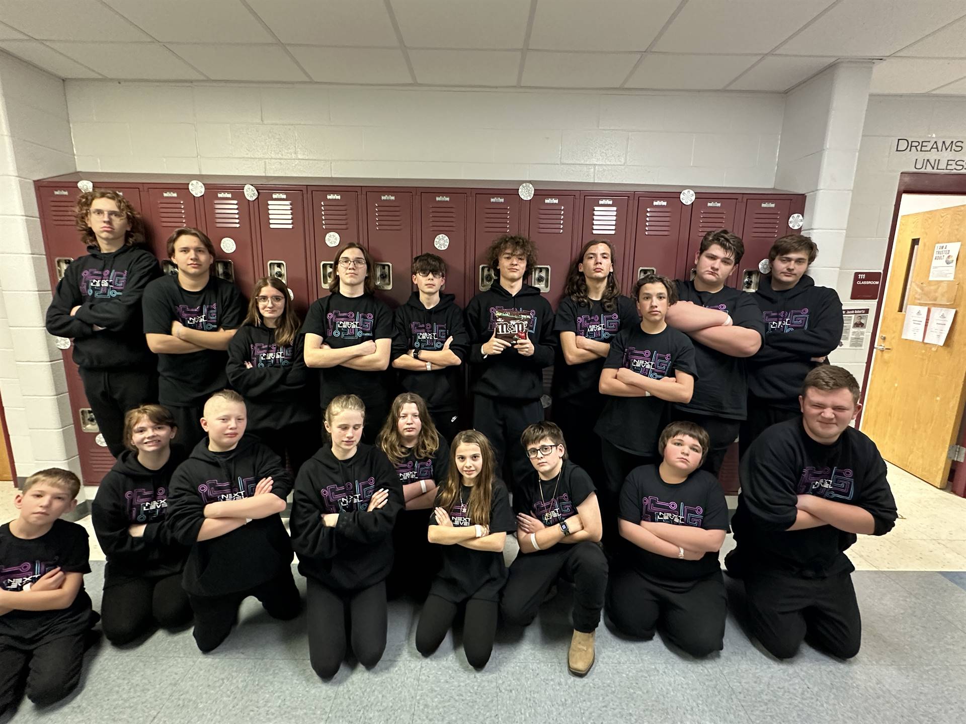 A group of students stands in a school hallway, wearing matching black sweatshirts and posing confidently with their arms crossed, in front of a row of lockers.