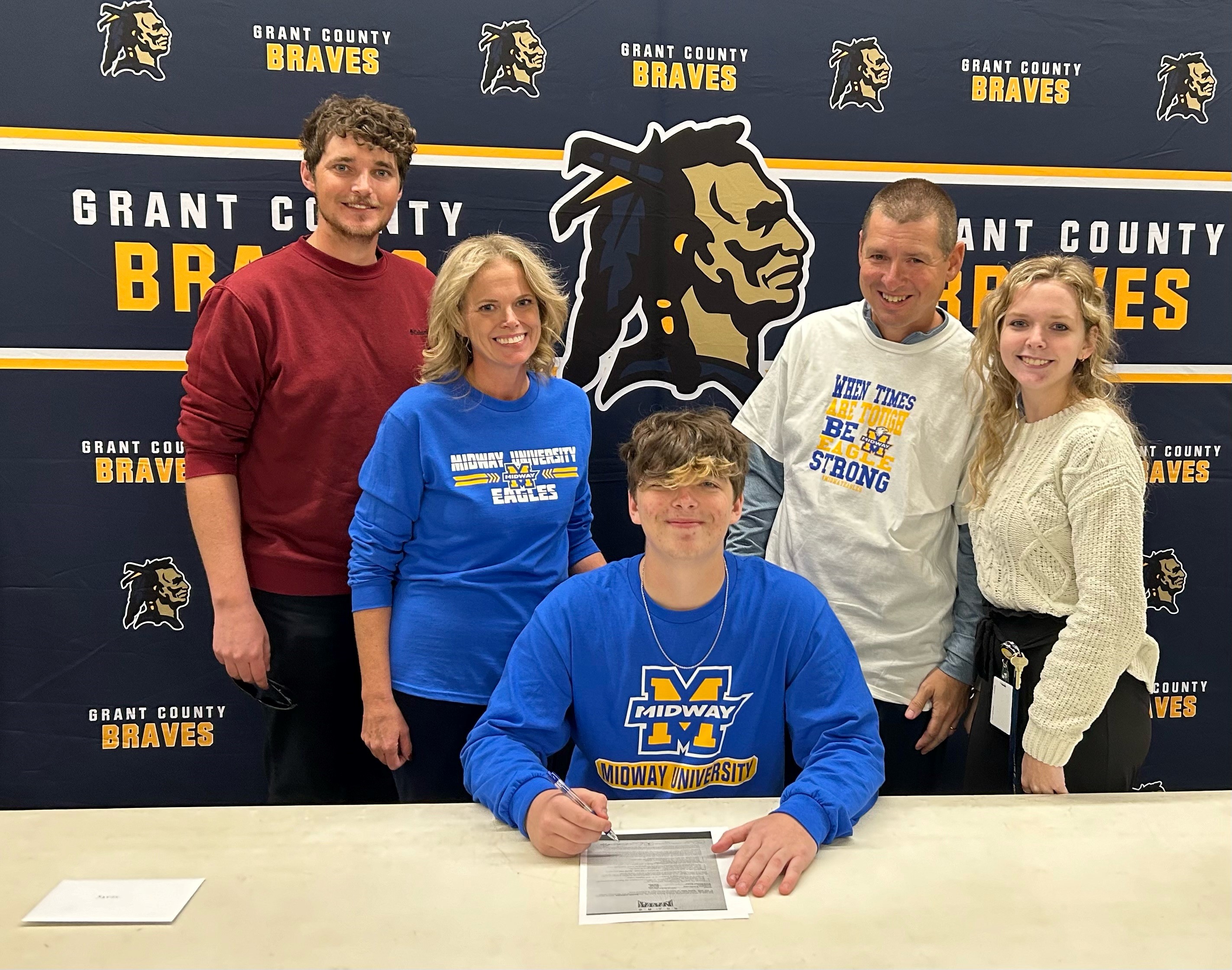 A group of five people, including a young man seated at a table signing a document, are gathered in front of a backdrop featuring the Grant County Braves logo, celebrating a significant event.