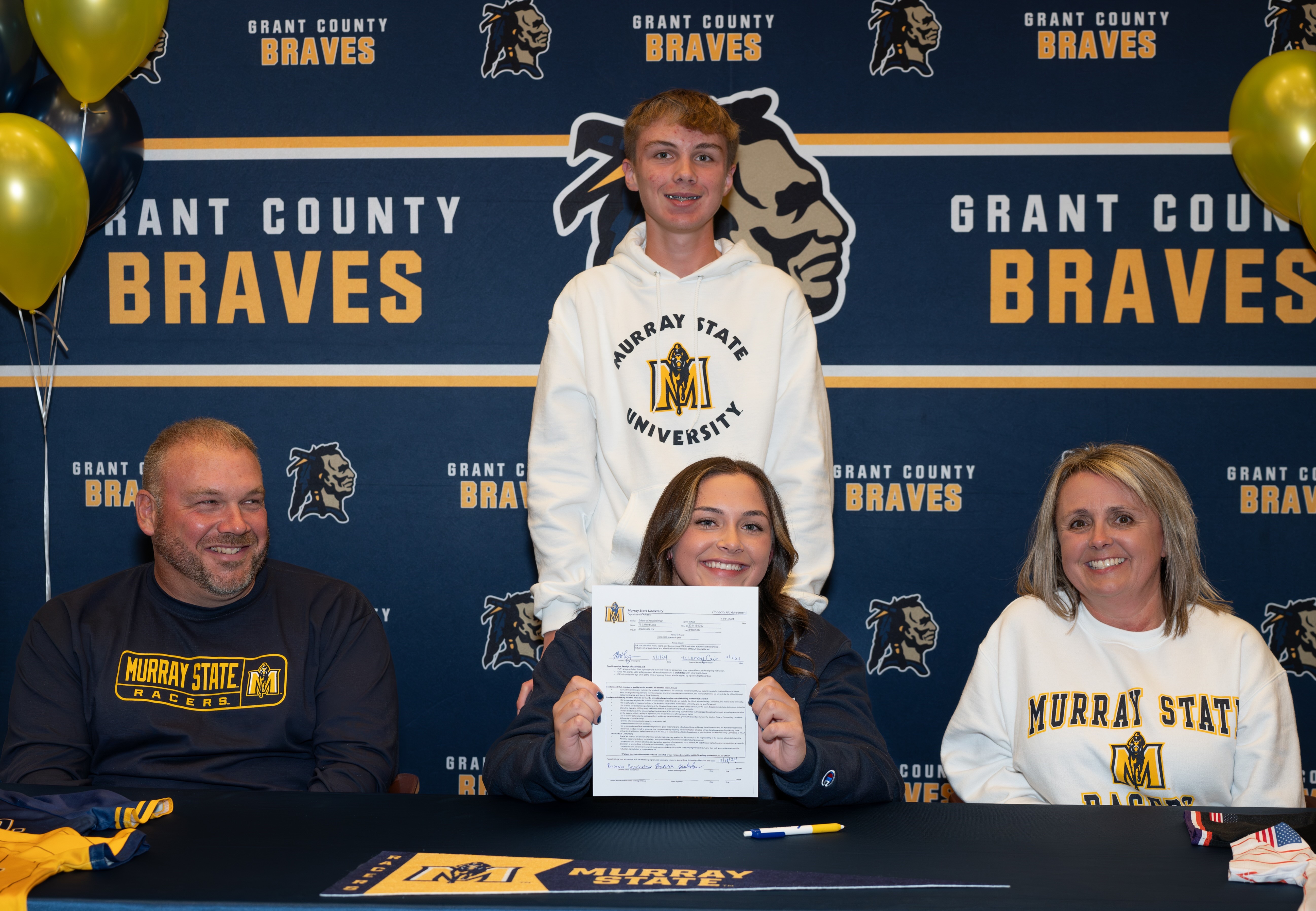 A group of four people, including a young man standing behind a seated woman holding a signed document, celebrate a signing event at a table decorated with balloons and a backdrop featuring the Grant County Braves logo.