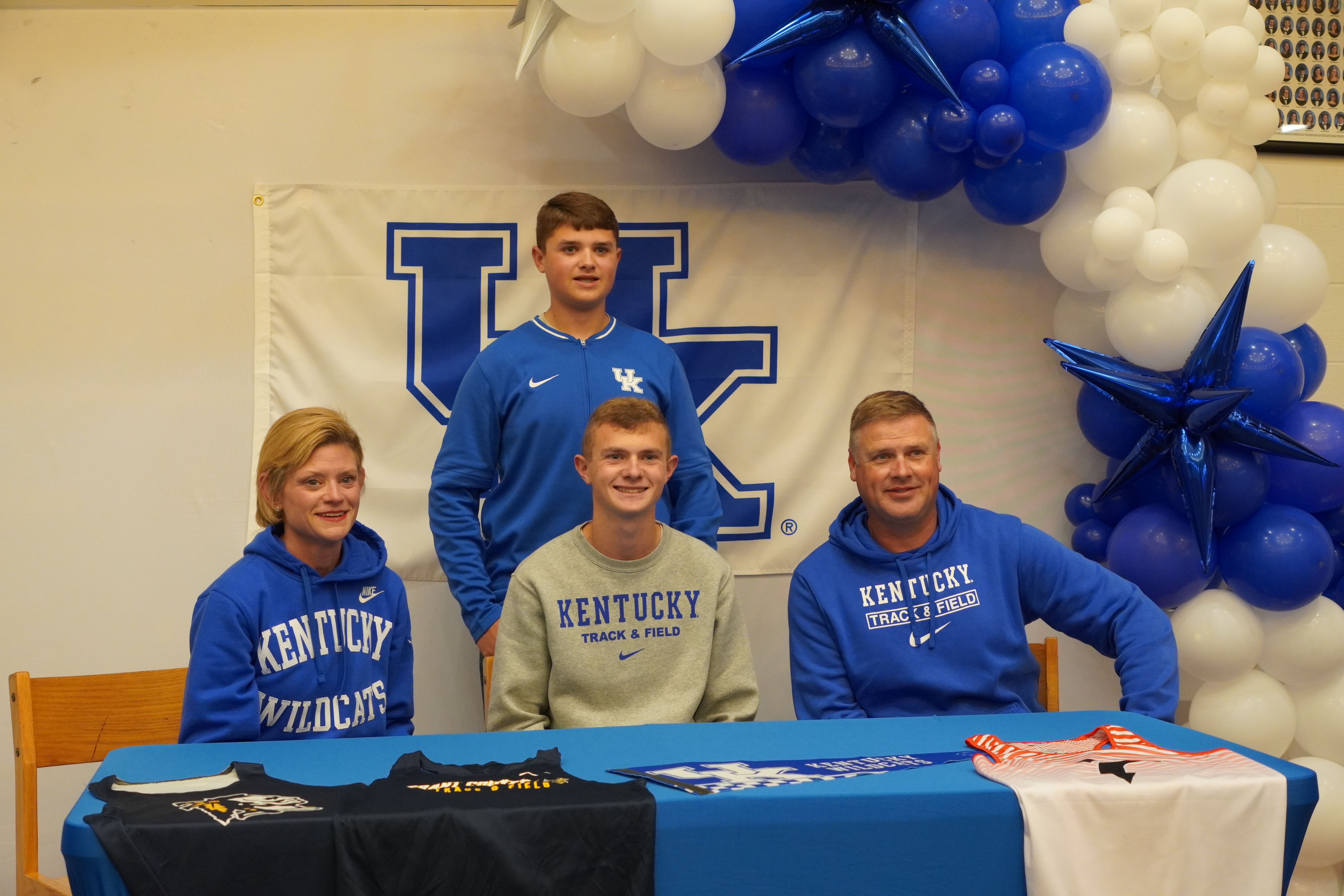Four students wearing Kentucky Wildcats apparel are seated at a table decorated with a blue and white balloon arch, celebrating a signing event.