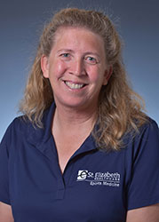 A smiling woman with curly blonde hair wearing a navy blue polo shirt with the logo of St. Elizabeth Healthcare.