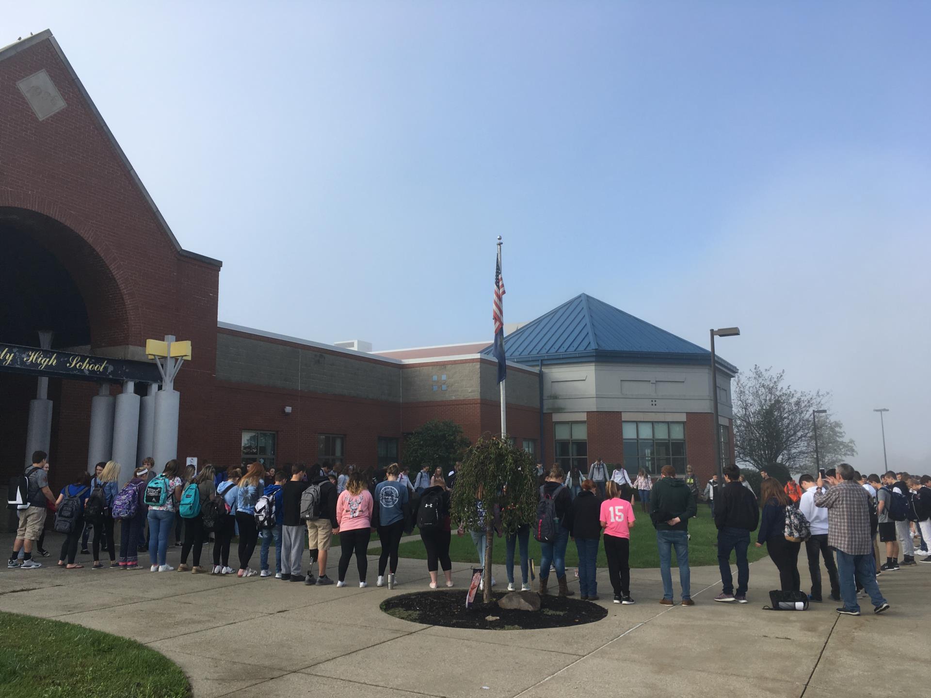 A long line of students waiting outside a school building on a foggy day, with a flagpole and greenery visible in the foreground.