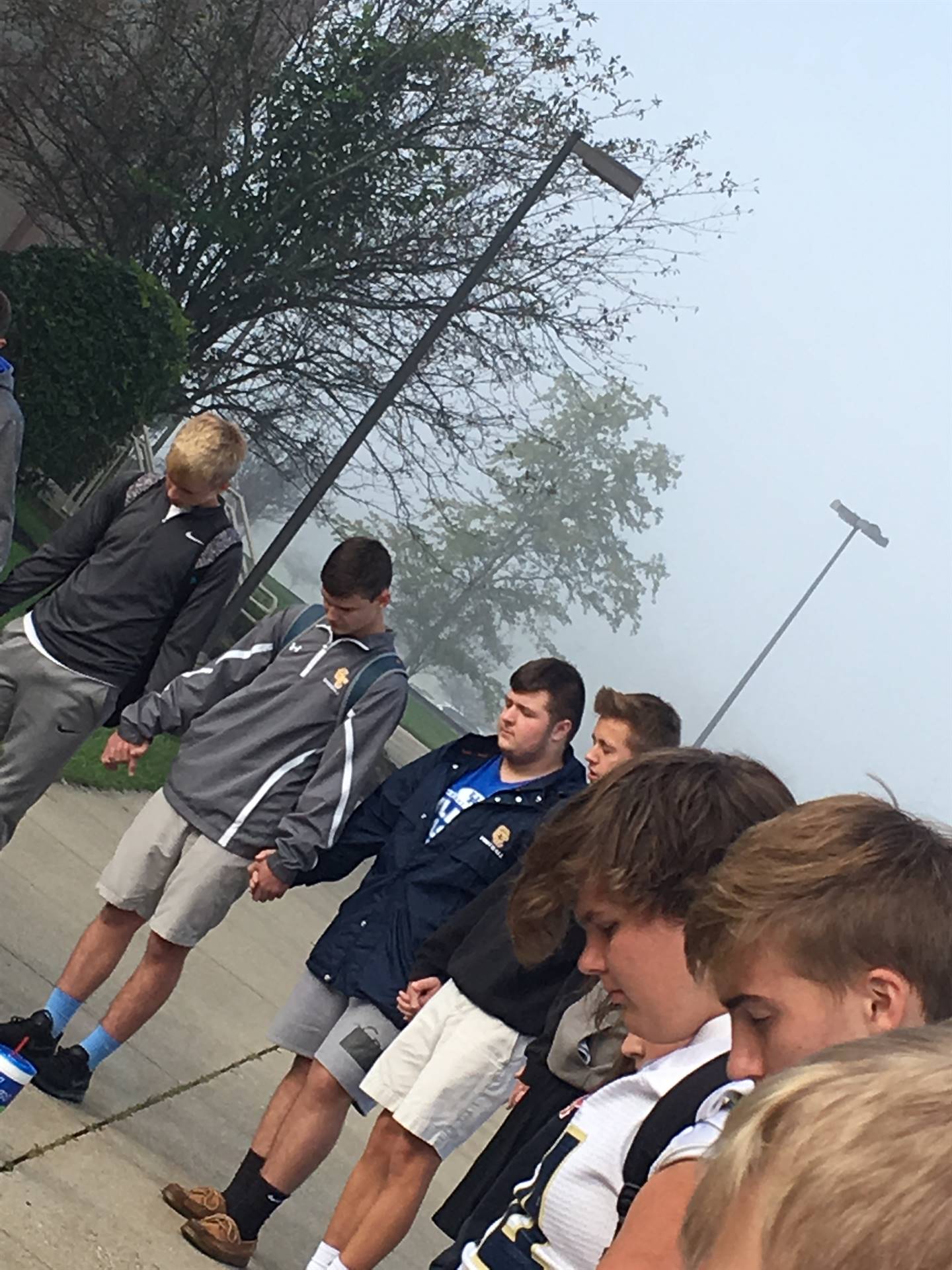 A group of young men in athletic attire stands outdoors, some facing away from the camera, with trees and a foggy background.