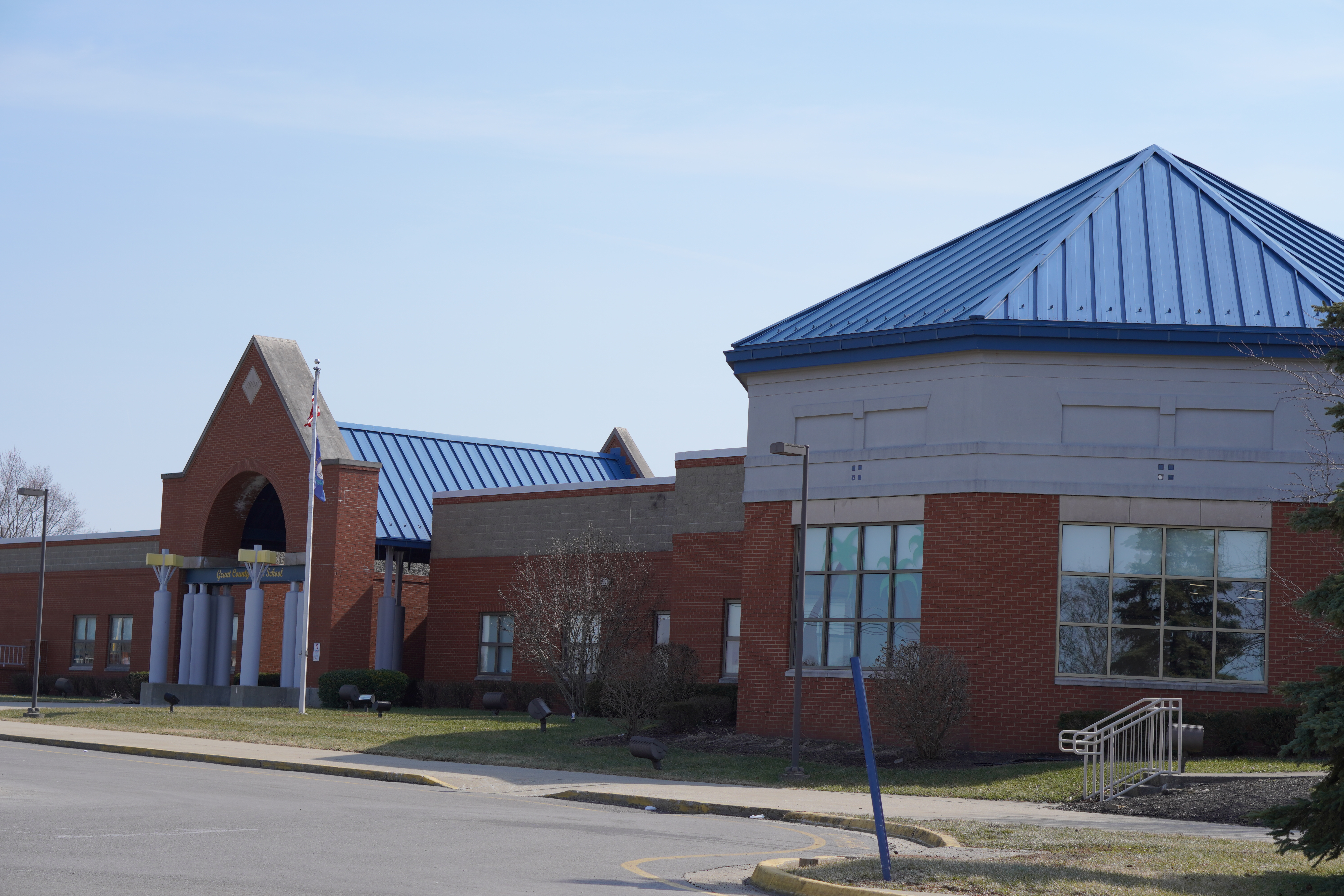A modern building with a blue metal roof and large windows, set against a clear sky, surrounded by a grassy area and a parking lot.
