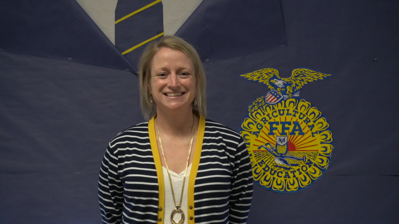 A smiling woman stands in front of a blue backdrop featuring the FFA emblem, wearing a striped shirt and a yellow necklace.