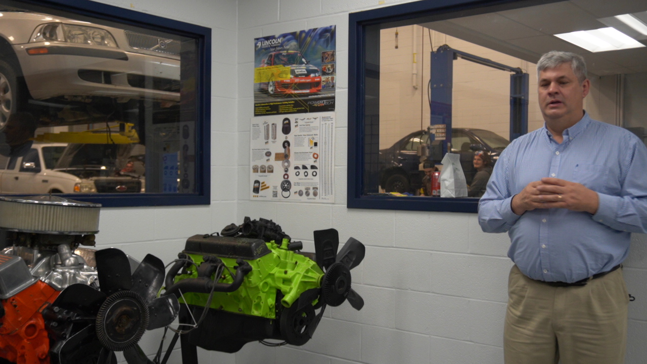 A man stands in a workshop next to a brightly colored engine, with vehicles visible through windows in the background and a poster on the wall featuring automotive diagrams.