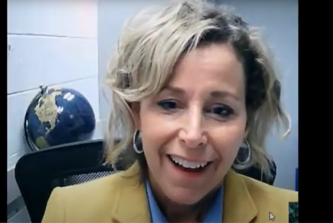 A woman with blonde, wavy hair smiles while sitting in front of a desk with a globe in the background, suggesting a professional video call setting.