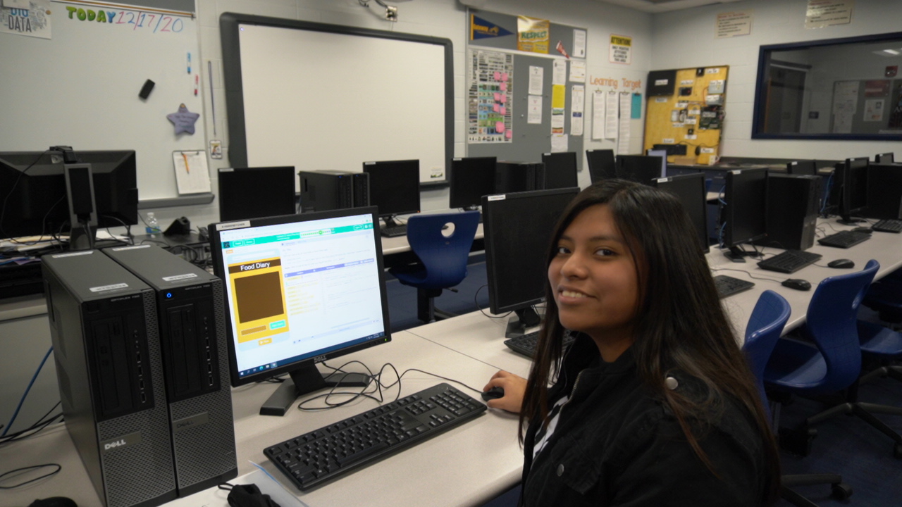 A student sits at a computer in a classroom filled with additional computers, smiling while working on a project.